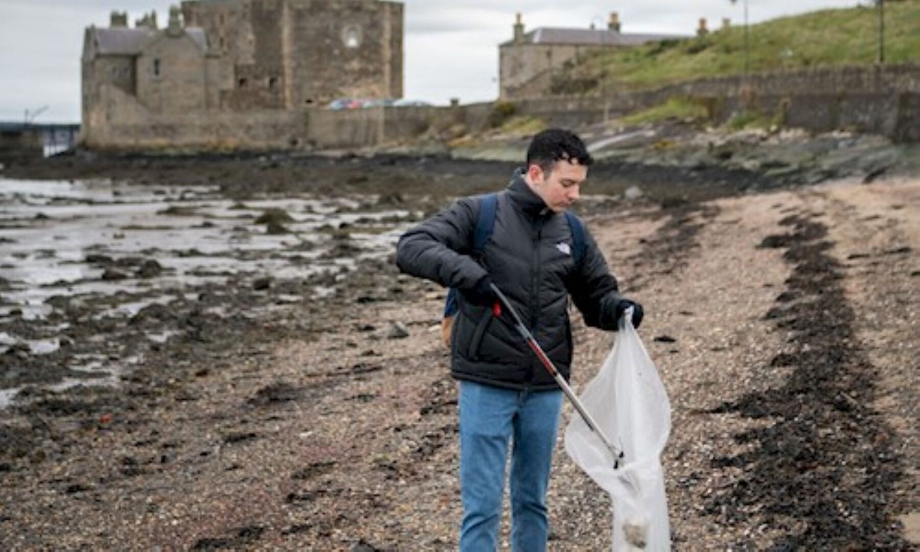 A man on a beach, with a castle in the background, putting rubbish in a bag.